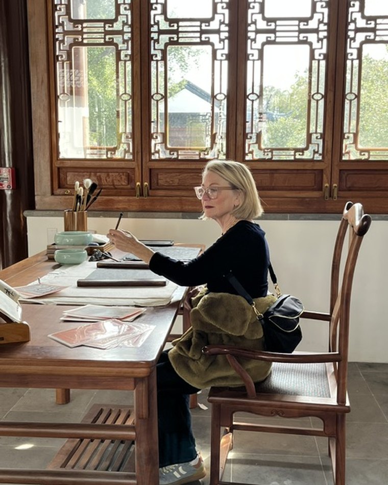 Laurie Schaefer seated at a long wooden table, brush in hand, working on paper by a window.