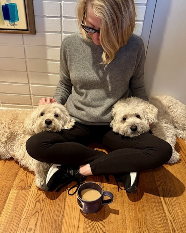 Laurie Schaefer sitting on the floor at home with her two dogs, Lucy and Buddy, one of her paintings on the wall behind.